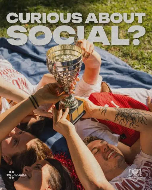 JMCC delegates lying on a blanket reach up to hold a silver trophy together. Curious About Social?