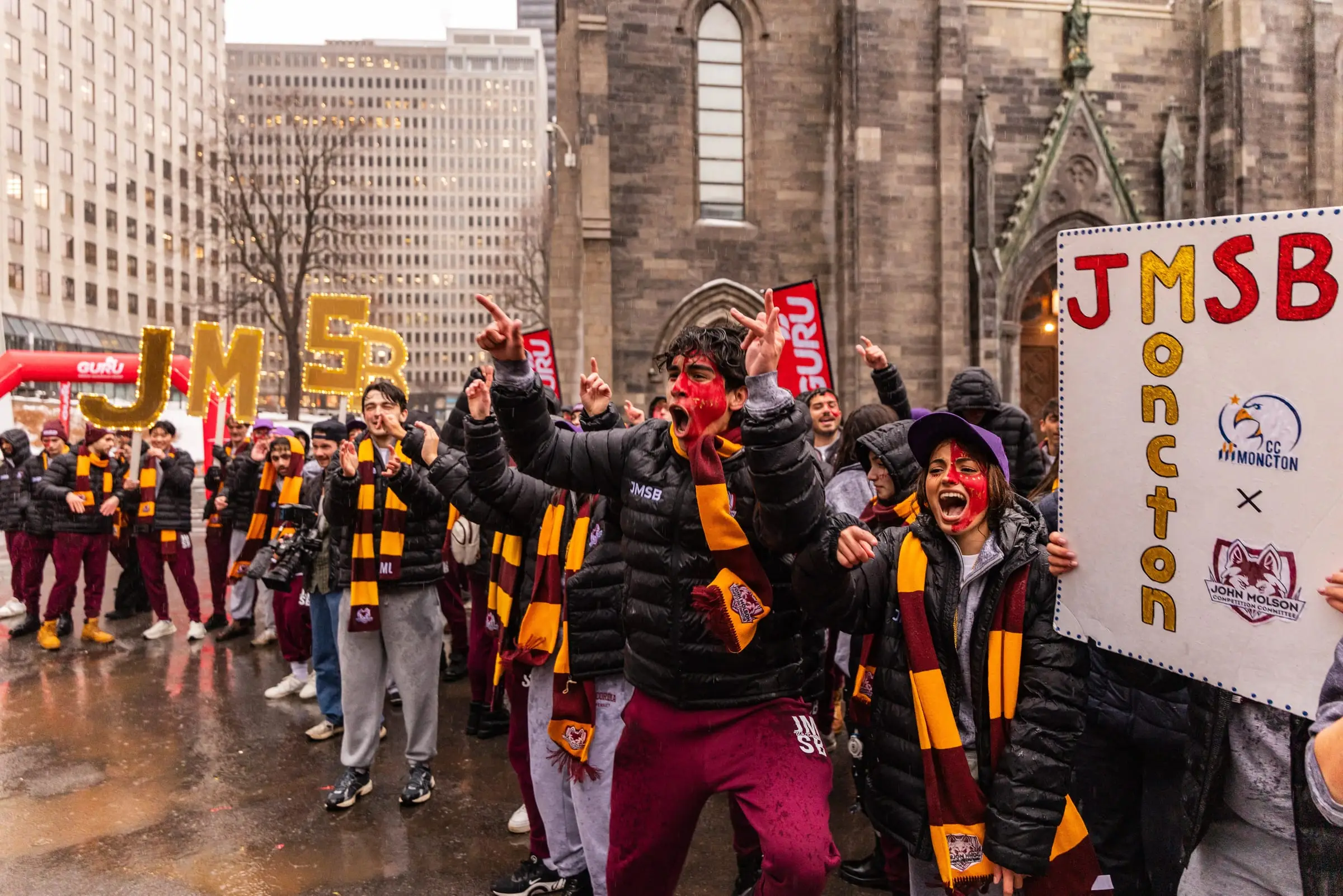The JMCC delegation marching and cheering in the rain behind lit JMSB letters at Jeux du Commerce 2026